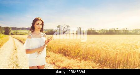 Beauté romantique fille appréciant la nature en plein air. Bonne jeune femme en short blanc tenant les oreilles sur la route près du champ de blé mûr doré. Banque D'Images