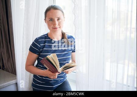 Jeune femme souriante debout à côté de la fenêtre tenant le livre dans l'isolement à la maison pendant la crise Covid-19. Auto-éducation en cas de pandémie 2020. Concept de sauver des vies chez soi. Copier l'espace Banque D'Images