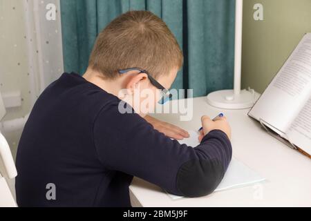 garçon dans des verres et une veste se trouve à une table et fait une tâche d'école Banque D'Images