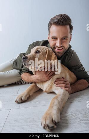 Mise au point sélective d'un homme souriant à la caméra tout en pétant Golden Retriever sur le sol sur fond gris Banque D'Images