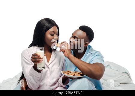 joyeux afro-américain homme nourrissant petite amie avec des biscuits dans le lit en pyjama isolé sur blanc Banque D'Images