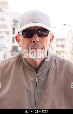 Décontracté habillé d'âge moyen ou homme âgé avec des lunettes de soleil noires, casquette blanche, portant coupe-vent et une petite barbe grise regardant directement dans le chamer Banque D'Images