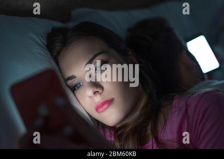 Concentration sélective de la femme addépendante utilisant un smartphone près de l'homme sur le lit la nuit Banque D'Images