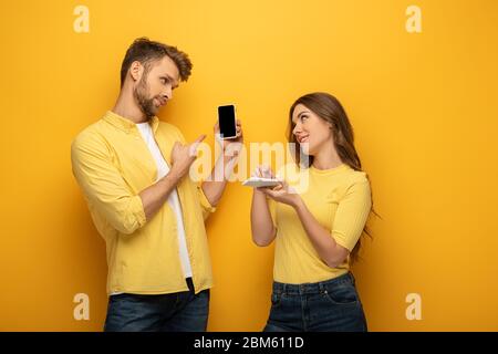 Jeune couple pointant sur un smartphone tout en regardant l'un l'autre sur fond jaune Banque D'Images