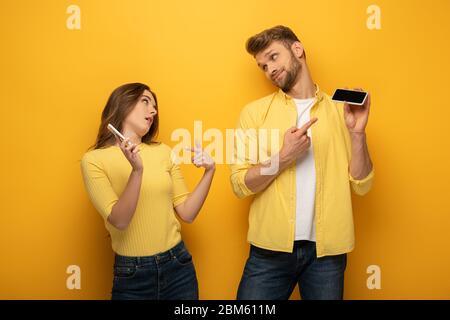 Couple pointant avec les doigts sur les smartphones et regardant l'un l'autre sur fond jaune Banque D'Images
