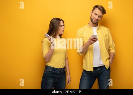 Une fille confuse regardant un ami souriant avec un smartphone sur fond jaune Banque D'Images
