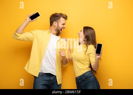 Jeune couple enthousiaste tenant un smartphone avec un écran vierge sur fond jaune Banque D'Images