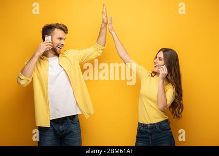 Couple joyeux haut cinq et souriant l'un à l'autre tout en parlant sur les smartphones sur fond jaune Banque D'Images