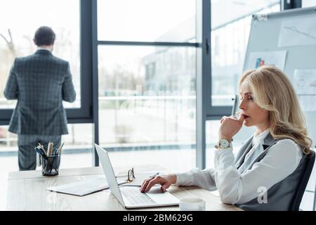 Objectif sélectif de femme d'affaires pensive utilisant un ordinateur portable alors que l'homme d'affaires se tenant près de la fenêtre dans le bureau Banque D'Images