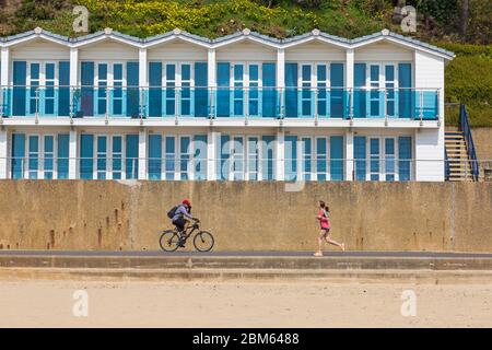 Poole, Dorset Royaume-Uni. 7 mai 2020. Temps au Royaume-Uni : belle journée chaude et ensoleillée avec des températures qui montent, prête pour le long week-end de vacances en banque. Les gens prennent leur exercice autorisé à la plage, la plupart en respectant les directives du coronavirus. Homme cycliste à vélo dans une direction et femme à jogging dans la direction opposée sur la promenade passant des cabanes de plage à Branksome Chine. Crédit : Carolyn Jenkins/Alay Live News Banque D'Images