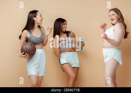 Sportswomen multiculturel avec ballon, bouteille de sport et haltères souriant et regardant l'un l'autre sur beige Banque D'Images