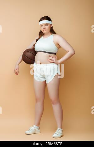 Plus taille sportswoman avec main sur la hanche et ballon regardant l'appareil photo sur beige Banque D'Images