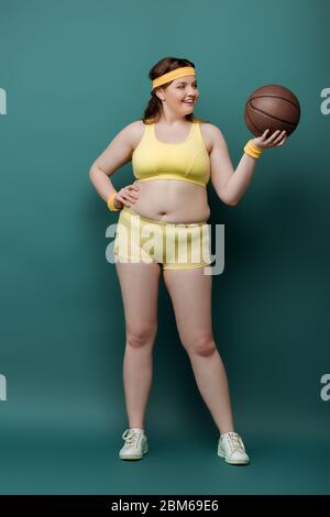 Grande taille sportswoman avec main sur la hanche regardant le ballon et souriant sur fond vert Banque D'Images