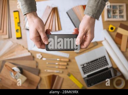 Décorateur accueil's hands holding a mobile phone à écran tactile, avec des outils de bureau, des ordinateurs portables et des échantillons de bois sur arrière-plan, Vue de dessus Banque D'Images