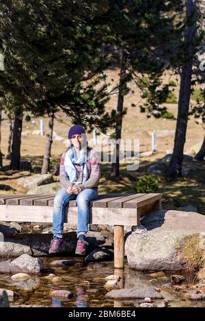 Femme portant un chapeau de laine assis sur un pont en bois au-dessus d'un ruisseau tout en prenant le soleil dans une forêt Banque D'Images