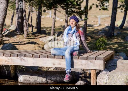 Femme portant un chapeau de laine assis sur un pont en bois au-dessus d'un ruisseau tout en prenant le soleil dans une forêt Banque D'Images