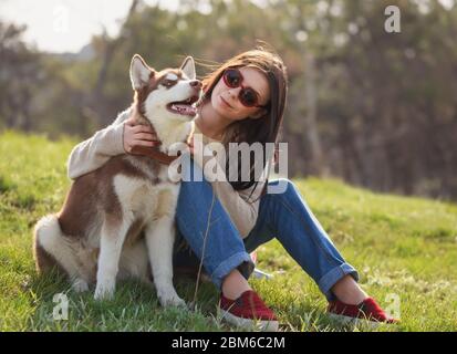 Jeune fille brune souriante assise avec son chien husky dans un parc vert à l'extérieur. Couple mignon et sympathique appréciant la nature. Jolie femme et molletonnée Banque D'Images