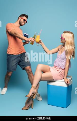 blonde, petite fille assise sur un réfrigérateur portable et un verre de jus d'orange avec un homme tenant une bouteille de bière, un passeport et des billets d'avion sur fond bleu Banque D'Images