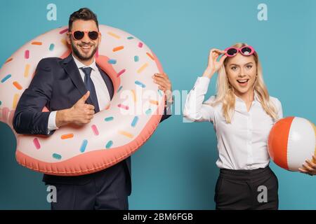 homme d'affaires heureux dans la bague de natation montrant le pouce vers le haut près de femme d'affaires souriante tenant le ballon gonflable sur fond bleu Banque D'Images