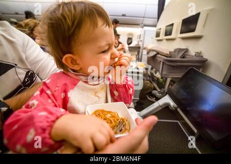 un petit tout-petit mignon est assis sur l'avion sur les genoux de ses parents et mange de délicieux repas spéciaux avec appétit. Banque D'Images