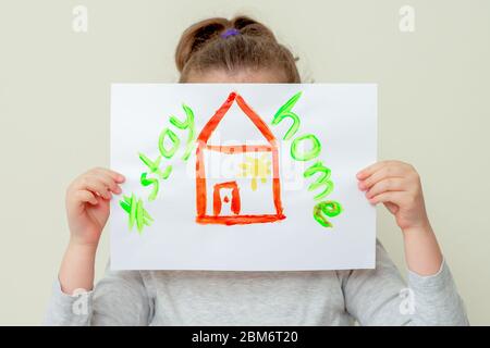 Les mains de l'enfant tenant une photo de maison rouge et les mots restent à la maison couvrant son visage. Enfants en quarantaine. Banque D'Images