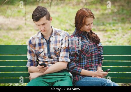 Un jeune couple assis dans un parc sur un banc regardant chacun dans leur téléphone mobile et regardant loin l'un de l'autre Banque D'Images