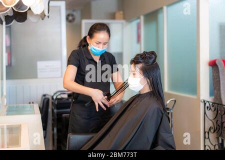 Jeune femme asiatique portant des masques pour se protéger contre Covid-19 pendant le coiffeur de tonte de cheveux noirs avec des ciseaux dans le salon de beauté. Banque D'Images