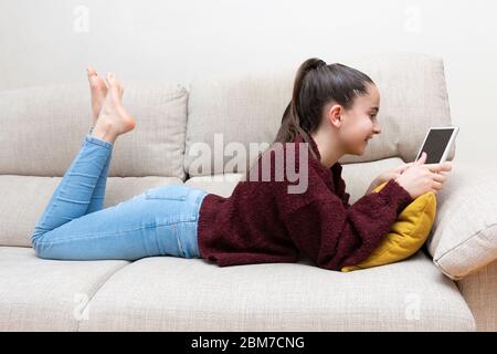 Profil d'une fille souriant pendant est étirée sur le canapé avec ses jambes soulevées à l'aide d'une tablette Banque D'Images