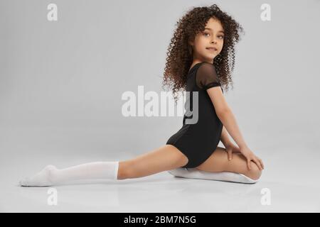 Vue latérale d'adorable fille souriante en noir sportswear et chaussettes mi-bas faisant demi-fendu, isolé sur fond gris studio. Petite gymnaste professionnelle féminine avec cheveux bouclés montrant la flexibilité. Banque D'Images