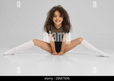 Fille souriante en chaussettes de sport noires et mi-bas avec cheveux bouclés pour plus de souplesse. Jeune femme gymnaste professionnelle assise sur le sol avec les jambes larges, isolée sur fond gris studio. Banque D'Images