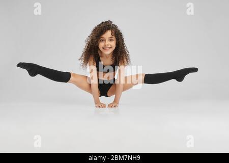 Petite femme professionnelle gymnaste faisant la fente, debout sur les bras au sol, isolé sur fond gris studio. Fille souriante en chaussettes de sport noires et mi-bas avec cheveux bouclés pour plus de souplesse. Banque D'Images