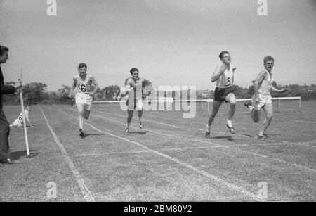 Journée des sports d'été au secondaire, Angleterre, c1960, des écoliers seniors se disputant une course de sprint et faisant un dernier effort avant de frapper la bande de finition tenue à côté de la piste d'herbe par un autre élève. Banque D'Images