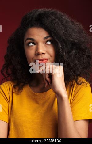 Image de la belle brunette africaine américaine avec la pensée de cheveux bouclés et touchant son menton isolé sur fond rouge Banque D'Images