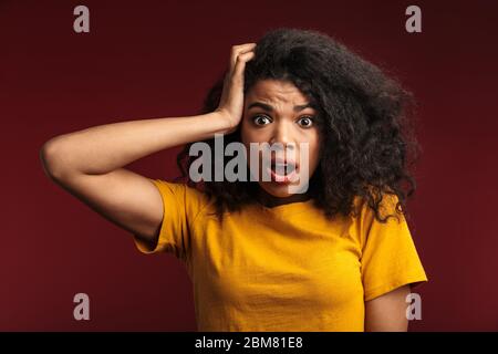 Image d'une jeune femme africaine confuse et émotive choquée posant isolée sur fond de mur rouge foncé. Banque D'Images