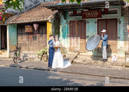 Hoi an, Vietnam - 12 avril 2018: Séance de photos et couple asiatique en tenue de mariage dans une rue de Hoi an avec un technicien tenant un réflecteur Banque D'Images