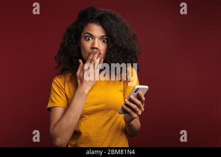 Image d'une femme afro-américaine brunette choquée avec des cheveux bouclés exprimant la peur tout en utilisant un téléphone cellulaire isolé sur fond rouge Banque D'Images