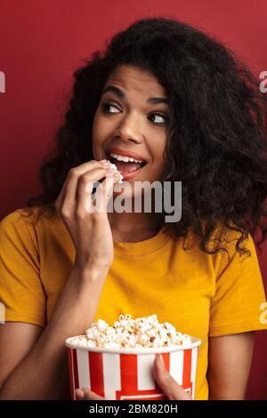 Image de la belle brunette africaine américaine avec les cheveux bouclés souriant et tenant le seau de pop-corn isolé sur fond rouge Banque D'Images