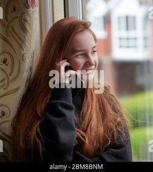 Ginger Ginger Ginger adolescente en noir décontracté sweat à capuche debout sur fond de fenêtre à la maison et de discuter avec des amis sur smartphone tout en regardant loin et de la sta Banque D'Images