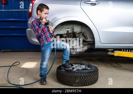 Un jeune garçon, un jeune travailleur automobile, fait un changement de pneu avec une clé pneumatique dans le garage d'une station de service. Un enfant apprend la changine mécanique Banque D'Images