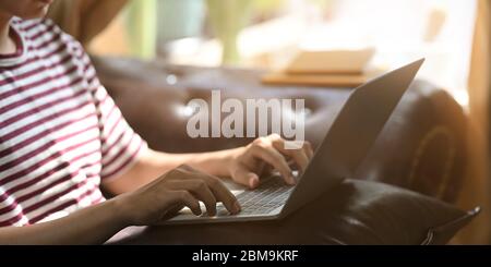 Image rognée d'un homme créatif utilisant/tapant sur un ordinateur portable qui met sur ses genoux tout en étant assis et se détendre sur le canapé en cuir confortable Banque D'Images