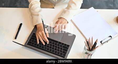 Image de dessus de la main de femme d'affaires dactylographiant sur ordinateur portable qui met sur le bureau blanc et entouré par un stylo, des crayons dans vase et de clipbo Banque D'Images