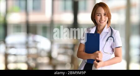 Belle femme travaillant comme personnel médical souriant et tenant un presse-papiers dans ses mains tout en se tenant sur l'hôpital flou comme arrière-plan. Banque D'Images