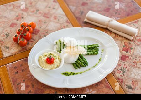 œuf poché aux asperges grillées, servi sur une assiette ovale blanche sous une croûte de fromage avec purée de pommes de terre et de tomates. table de salle à manger vintage Banque D'Images