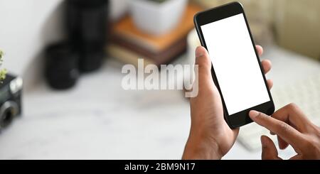 Image rognée des mains d'un homme intelligent tenant un smartphone noir court avec un écran blanc blanc sur son bureau blanc comme arrière-plan. Banque D'Images