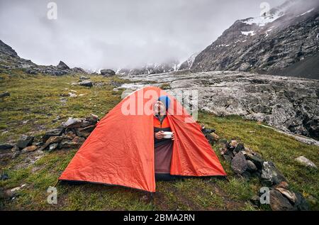 Happy tourist est en train de manger son petit-déjeuner à orange tente dans la montagne brumeuse matinée pluvieuse au parc national à Karakol, Kirghizistan Banque D'Images