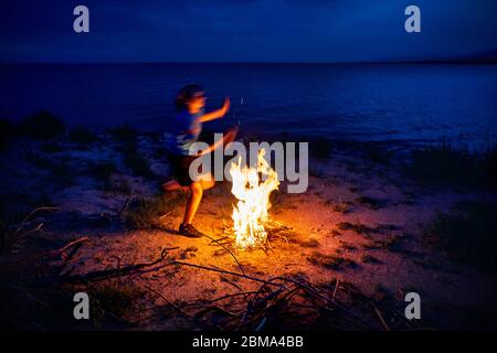 Jeune homme saute au-dessus du feu de camp sur la plage près du lac au crépuscule Banque D'Images