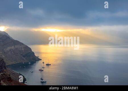 Grèce. De lourds nuages au-dessus de la caldeira de Santorini. Des rayons de soleil éclairent la baie et plusieurs yachts ancrés Banque D'Images