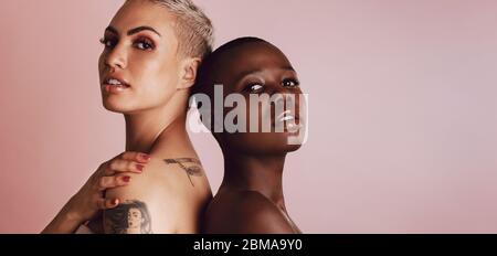 Divers modèles féminins fiables, debout dos à dos et regardant l'appareil photo. Deux femmes multiethniques se tenant ensemble sur fond beige. Banque D'Images