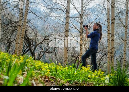 Un photographe de l'homme se tient sur la piste dans la forêt de bouleau de printemps. Le gars tient une caméra dans sa main. Vue de l'arrière. Herbe jeune fraîche et friche Banque D'Images