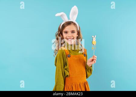 Photo de joyeuse fille de curly dans les oreilles de lapin jouet tenant le lapin jouet et souriant isolé sur fond bleu Banque D'Images